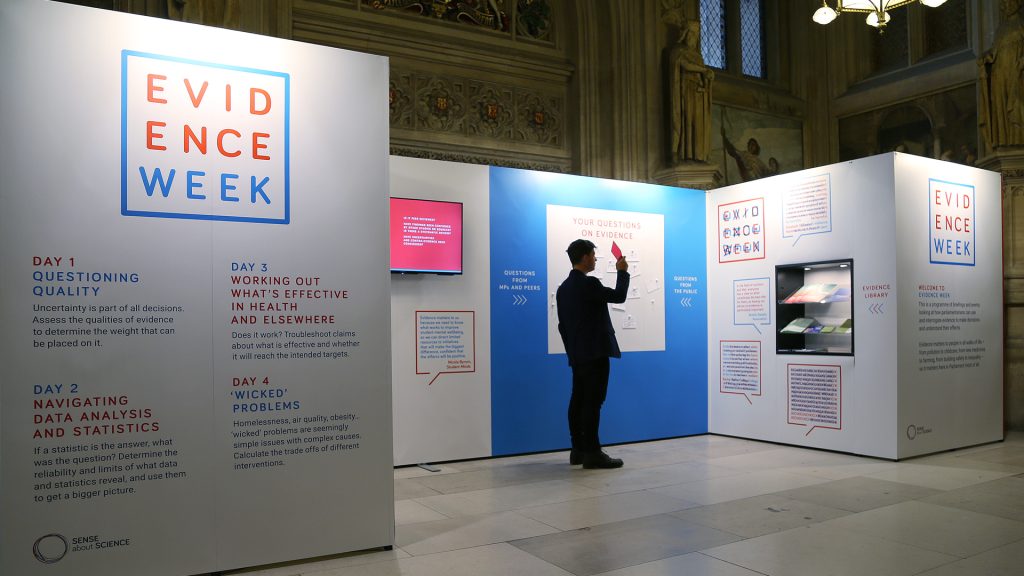 A person standing in front of the Evidence Week exhibition stand holding a red viewing glass in the UK Parliament.