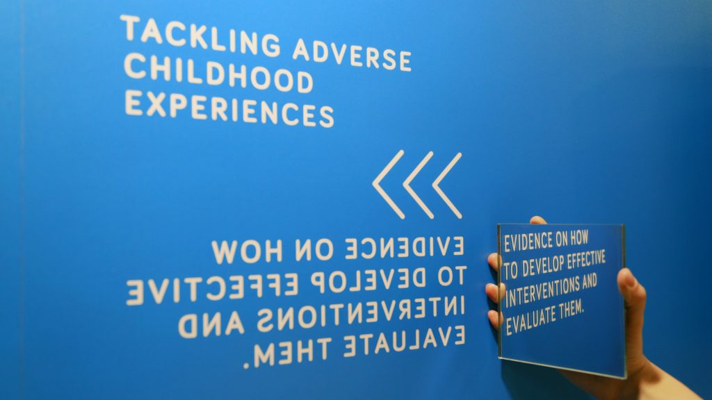 A hand holding a square shaped mirror agains the blue exhibition stand to reveal a white text message that has been printed in reversed direction.