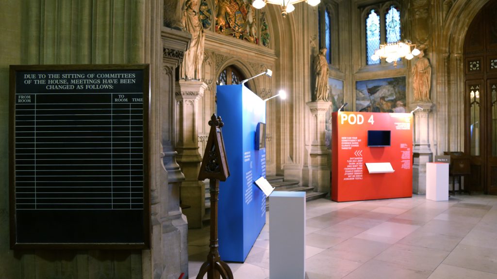 Photograph showing half of the Upper Waiting Hall of the UK Parliament with two exhibition stands and two pillars. 