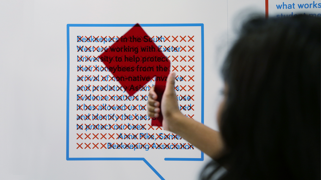 A person holds a red viewing glass up to the Evidence Week exhibition stand, revealing hidden text.