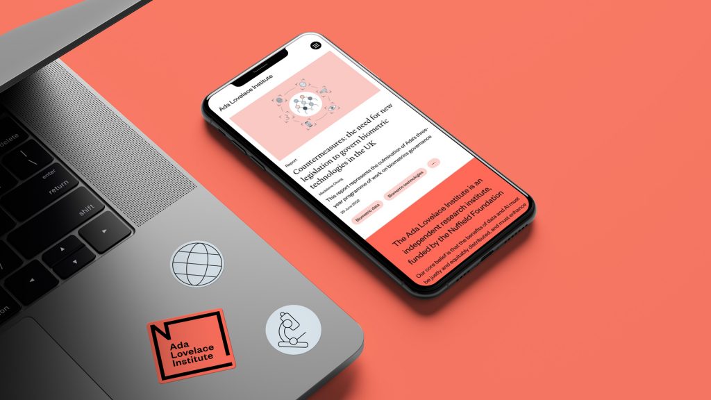 The Ada Lovelace website shown on a mobile device next to a corner of a laptop with stickers of the Ada Lovelace logo and supporting illustrations.