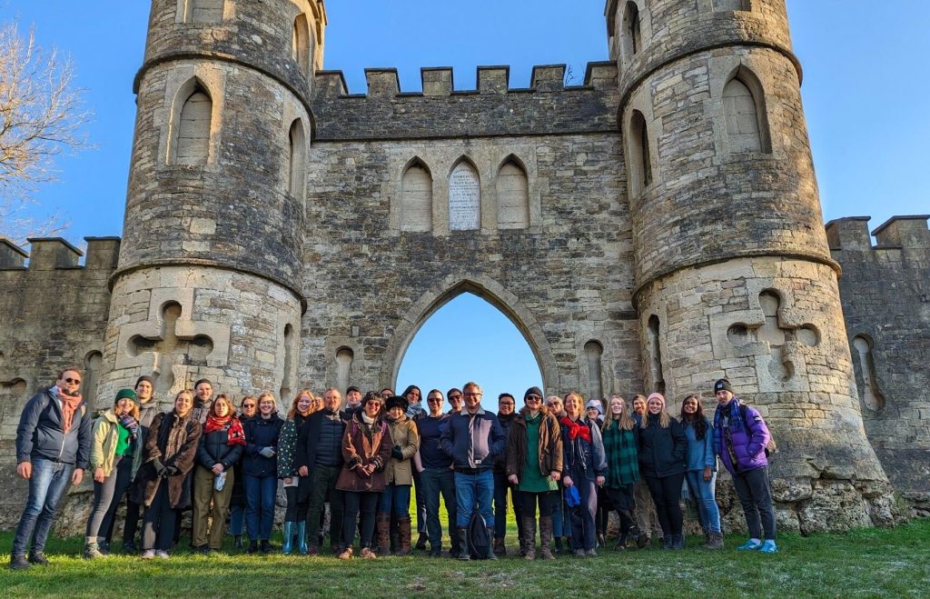 Outdoor photo of around 30 people stood in a line, dressed in warm clothes in the sunshine, in front of a ruined old gatehouse.